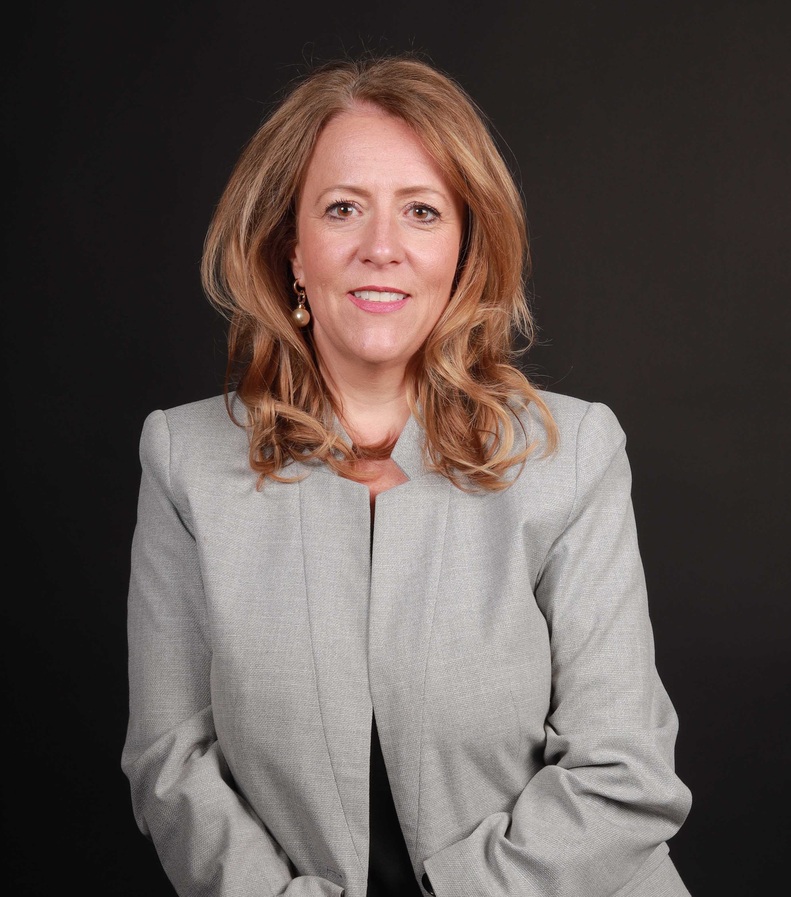 Maxi wearing grey suit with her hair down smiling at the camera against a black background