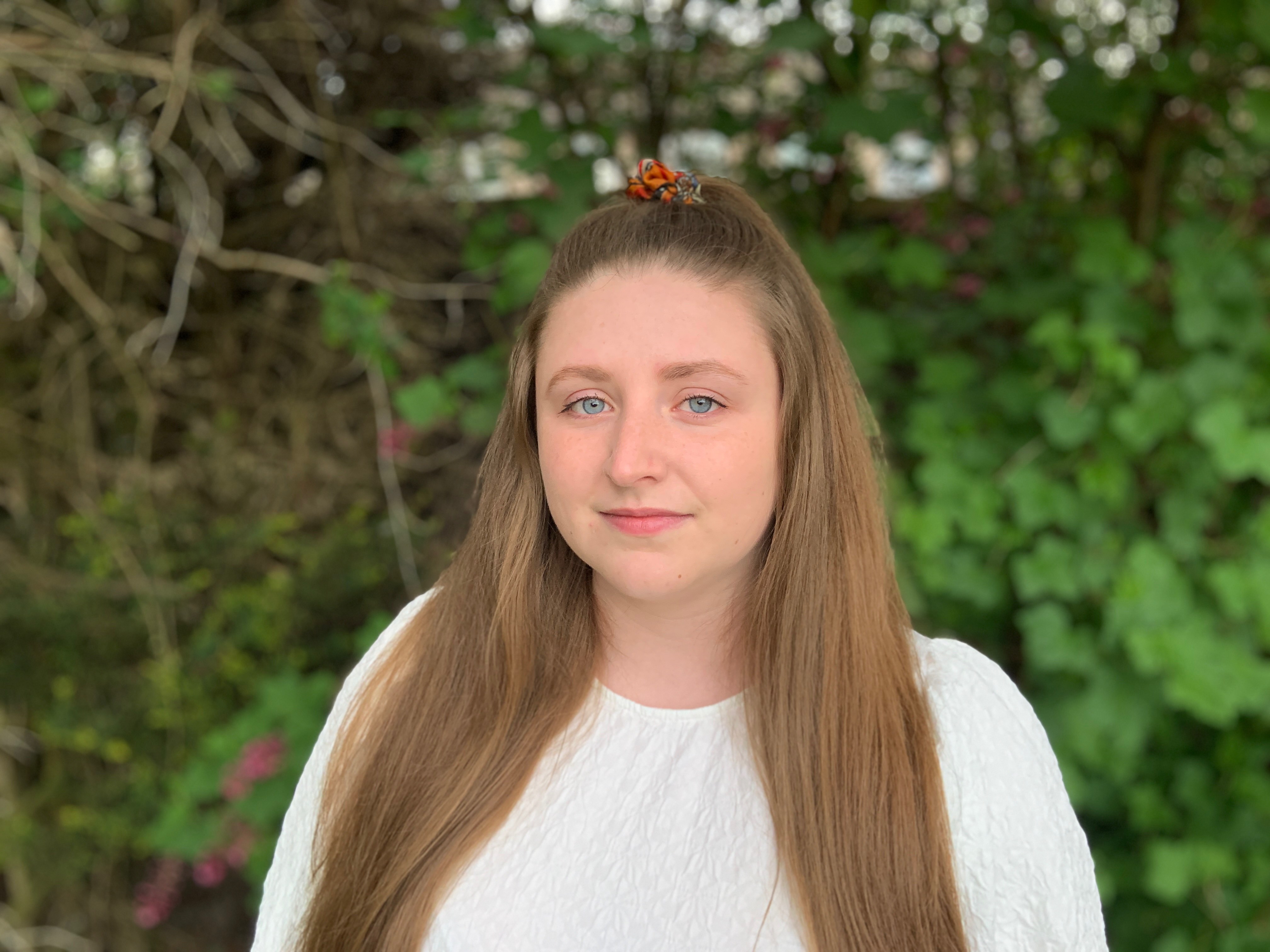 Photo of Elizabeth Simon. She is wearing a white jumper, has long mid-brown hair, and is standing in front of a hedge.