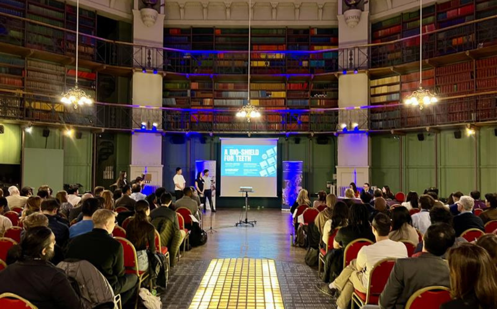 An image of people standing on stage presenting to a large group of people sat on chairs with a walkway in between